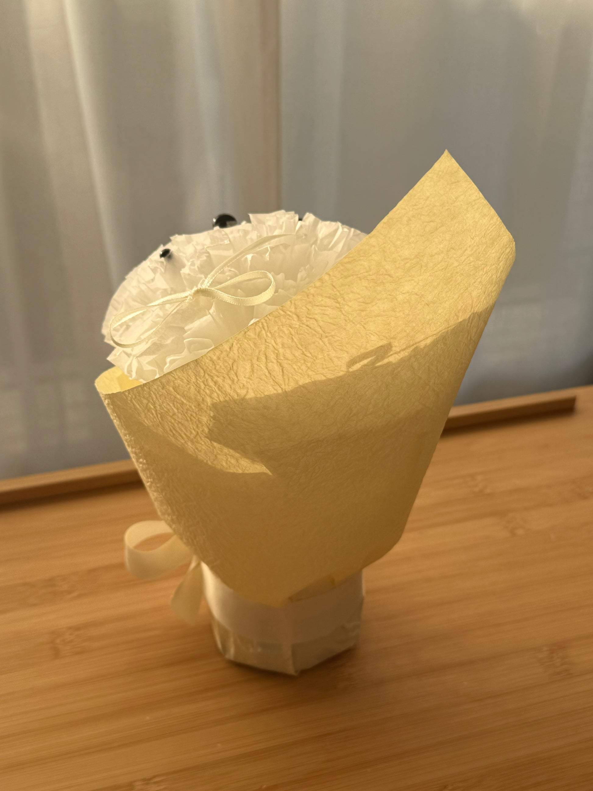 A small bouquet wrapped in textured cream paper and tied with a ribbon, standing on a wooden surface with a soft indoor background.